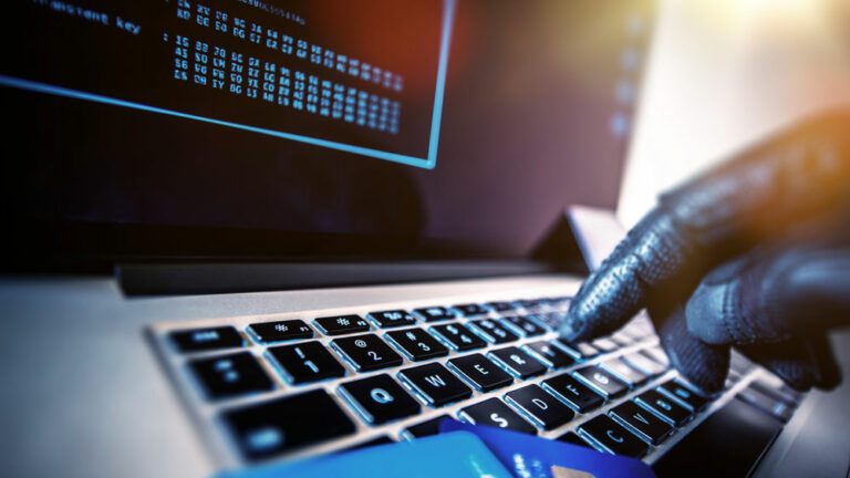 Gloved hand typing on a laptop with a menacing look, holding debit cards. Cyberfraud concept.